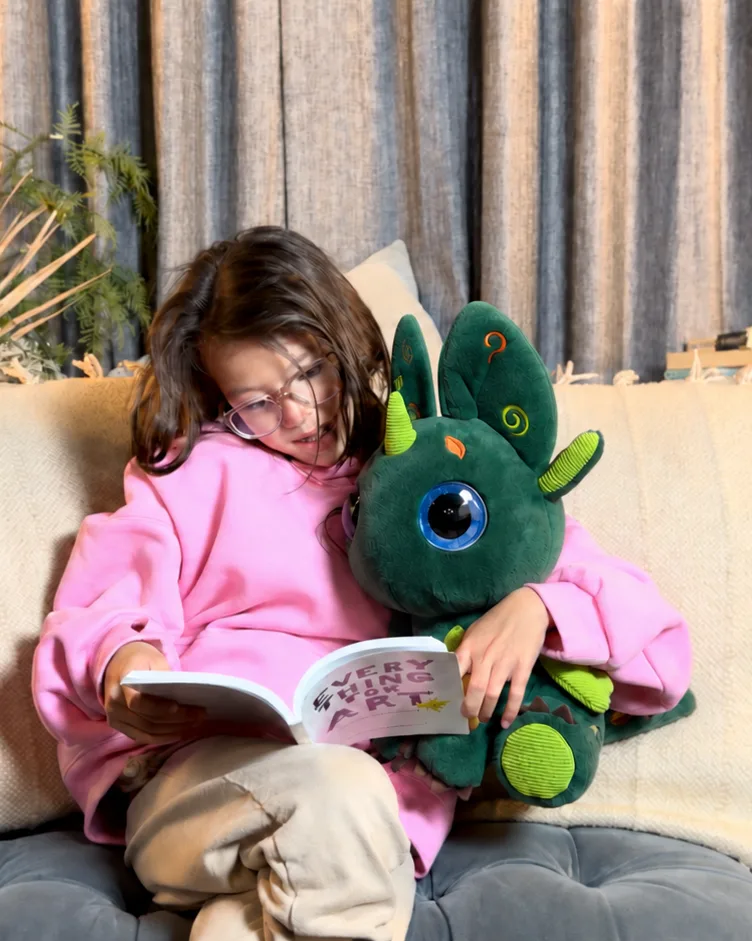 A girl in a pink hoodie reading a picture book on a sofa, with Bloom held in her lap.