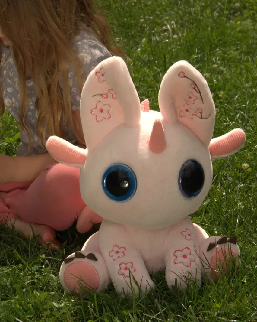A child sits in the grass beside Bloom, SkyBloom's plush companion in Cherry Blossom Pink.