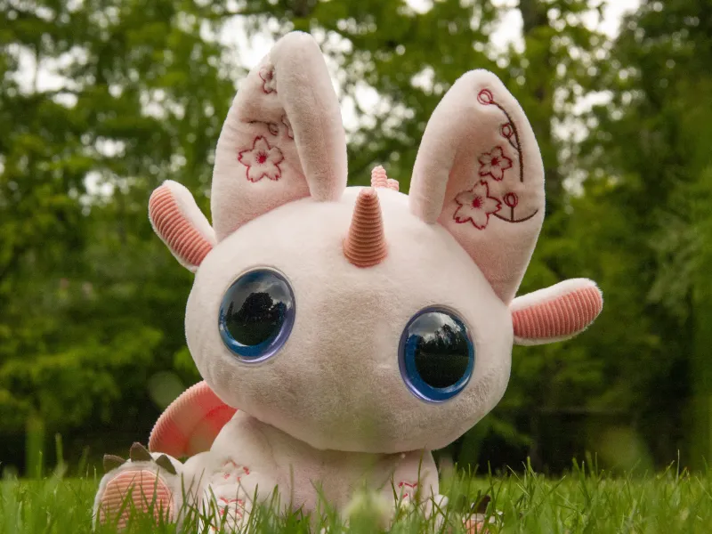 Bloom, a pink plush companion with tall ears, sitting in grass with trees behind.